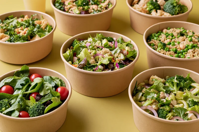 Multiple kraft paper bowls filled with fresh salads arranged on a yellow surface