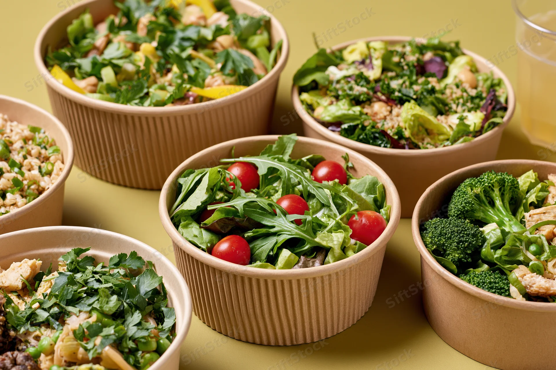 Multiple kraft paper bowls filled with fresh salads and vegetables arranged on a yellow surface