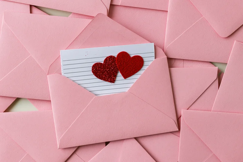 Open pink envelope revealing a lined card with two red glitter hearts inside, surrounded by stacked pink envelopes