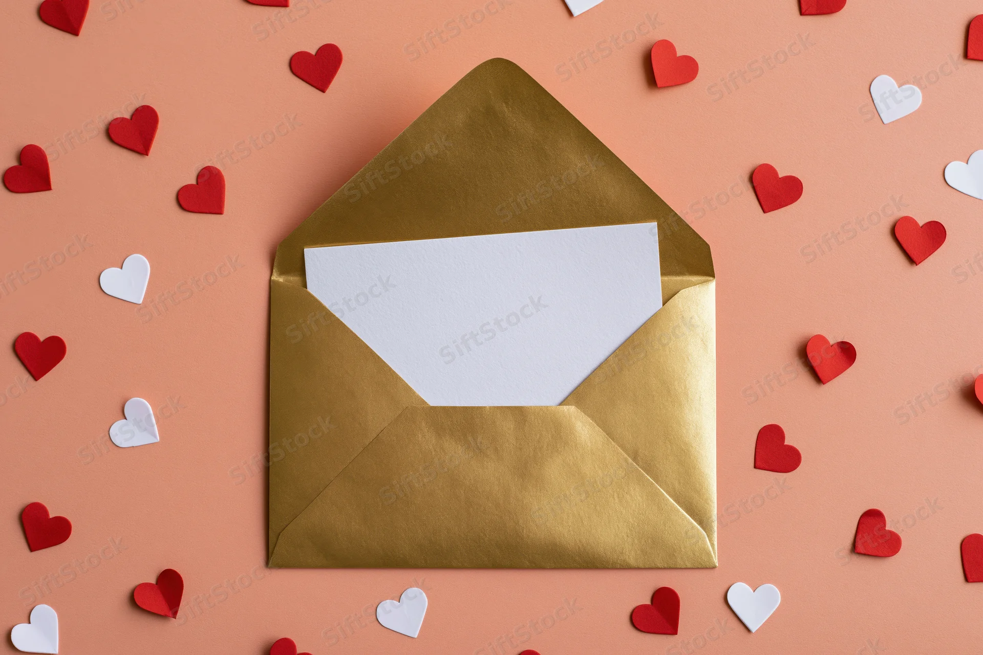 Open gold metallic envelope with blank white card surrounded by red and white paper hearts on peach background