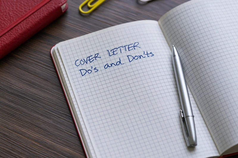 Open grid-lined notebook with handwritten cover letter do's and don'ts on dark wooden desk with pen and red notebook nearby