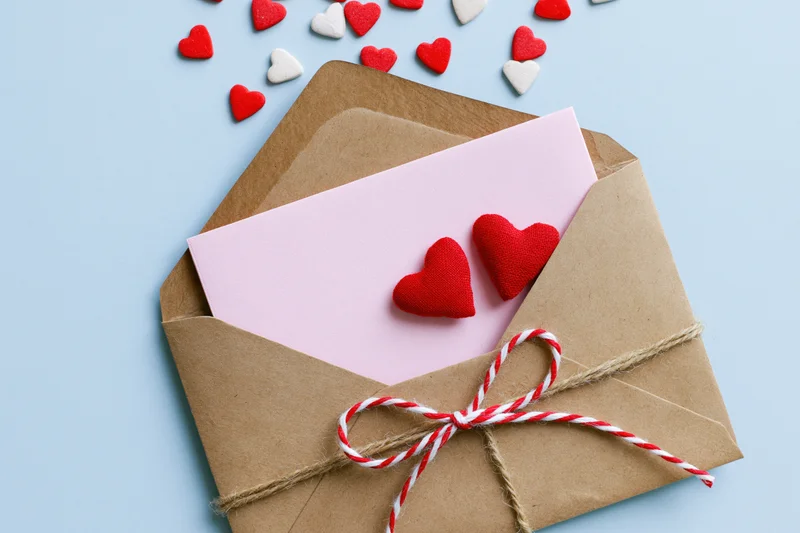 Open kraft envelope with pink note and red hearts tied with twine on light blue background