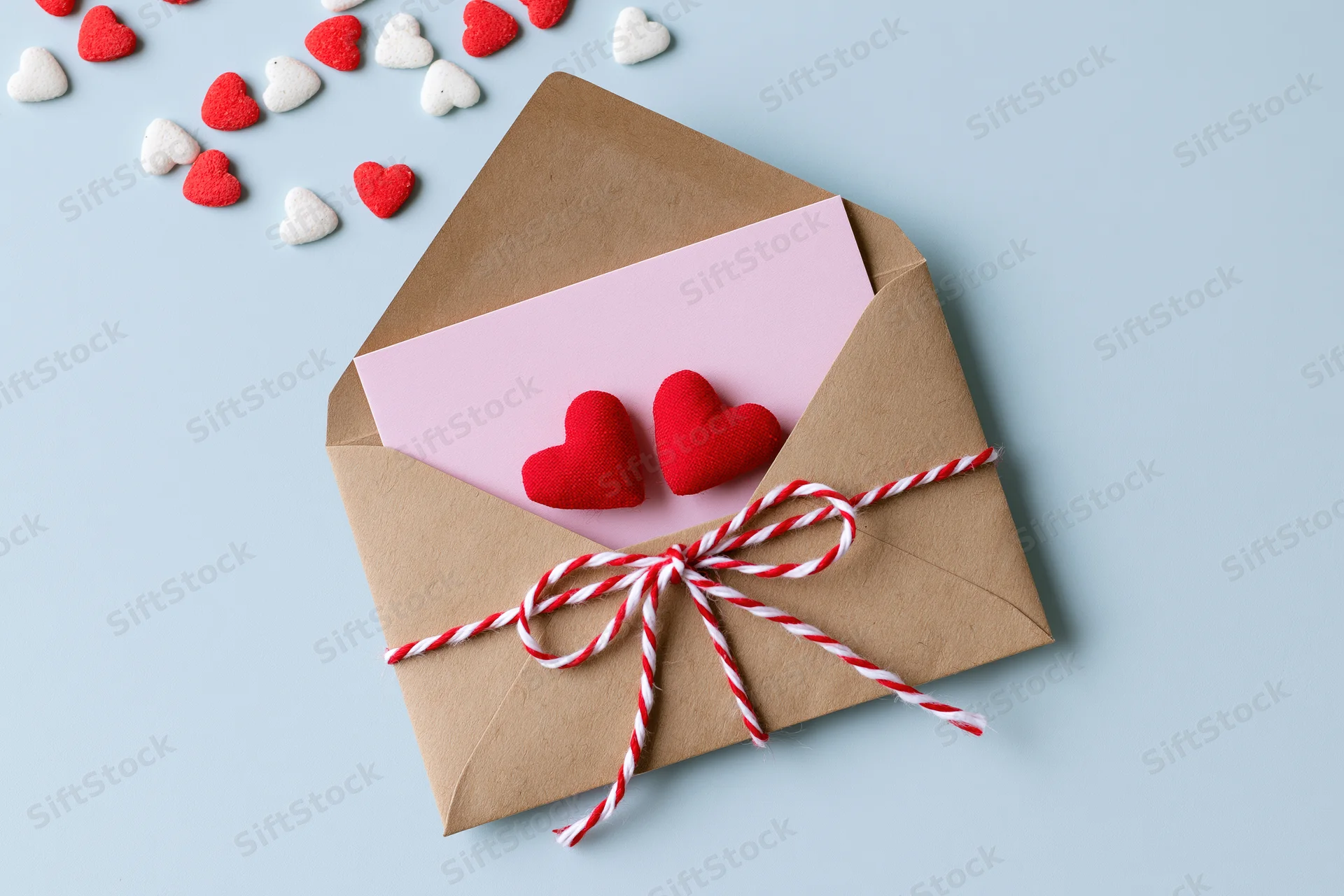Open kraft envelope with pink note and red hearts tied with striped ribbon on light blue background