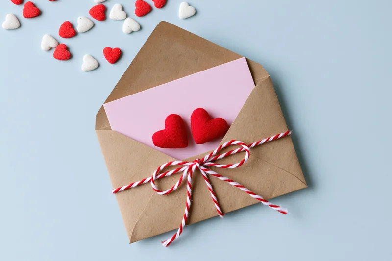 Open kraft envelope with pink note and red hearts tied with striped ribbon on light blue background