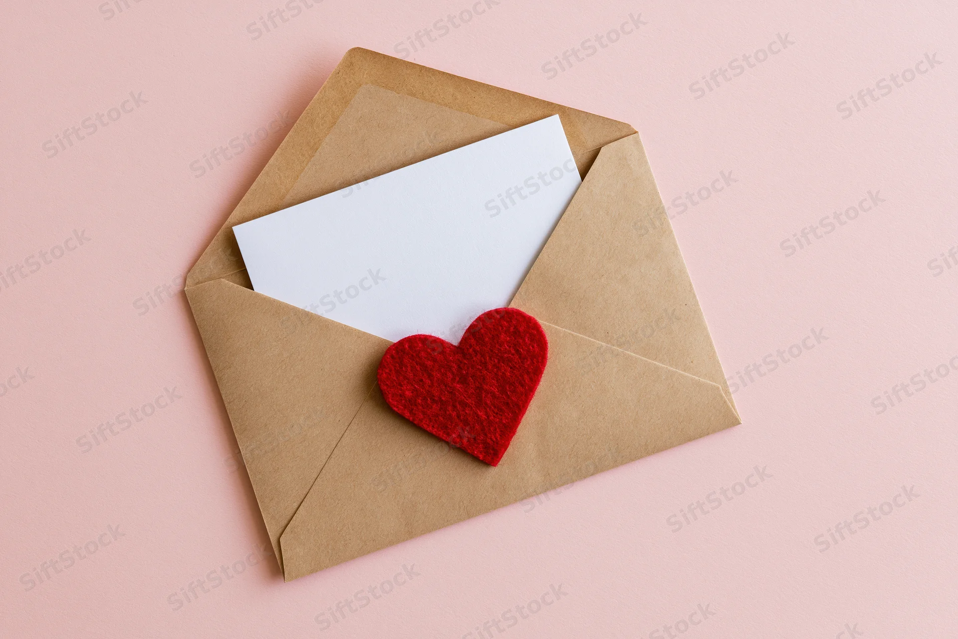 Open kraft envelope with red felt heart and white card on pink background