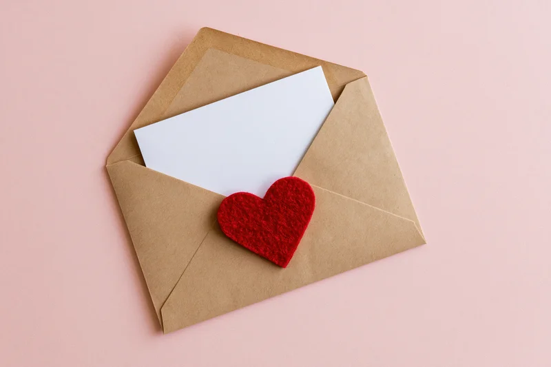 Open kraft envelope with red felt heart and white card on pink background