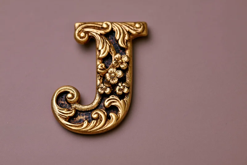 Ornate gold metal letter j with embossed floral scrollwork on a muted purple background