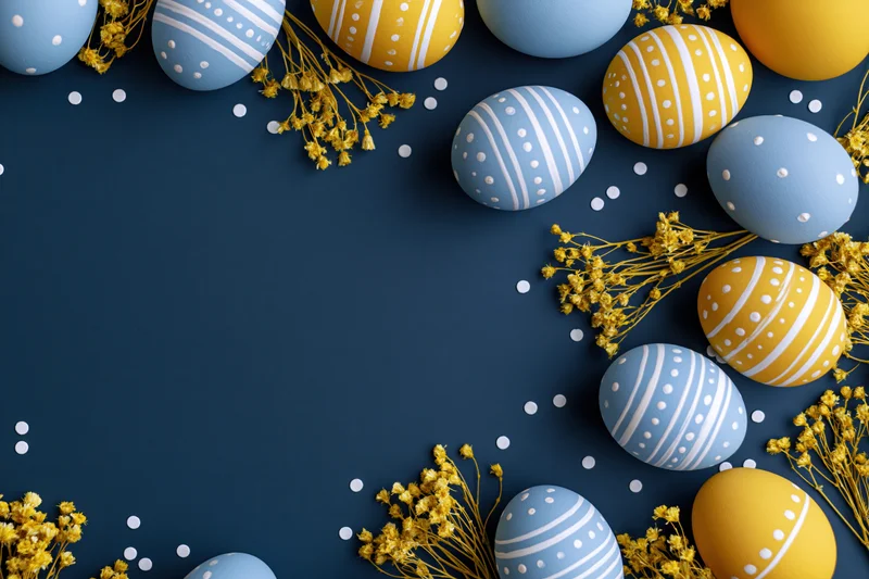 Painted easter eggs in blue and yellow with floral sprigs arranged in a border on navy background with copyspace