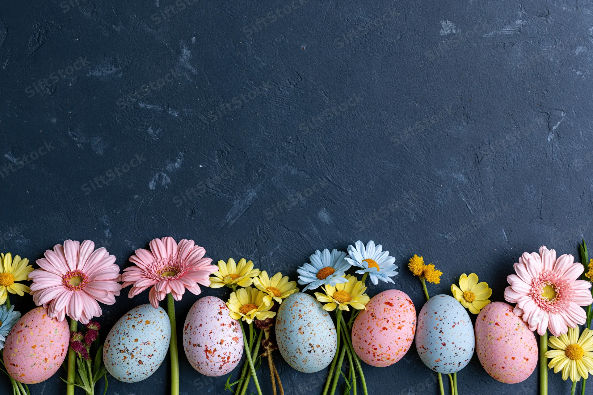 Pastel speckled easter eggs and spring daisies arranged in a row along a dark textured surface with copyspace above