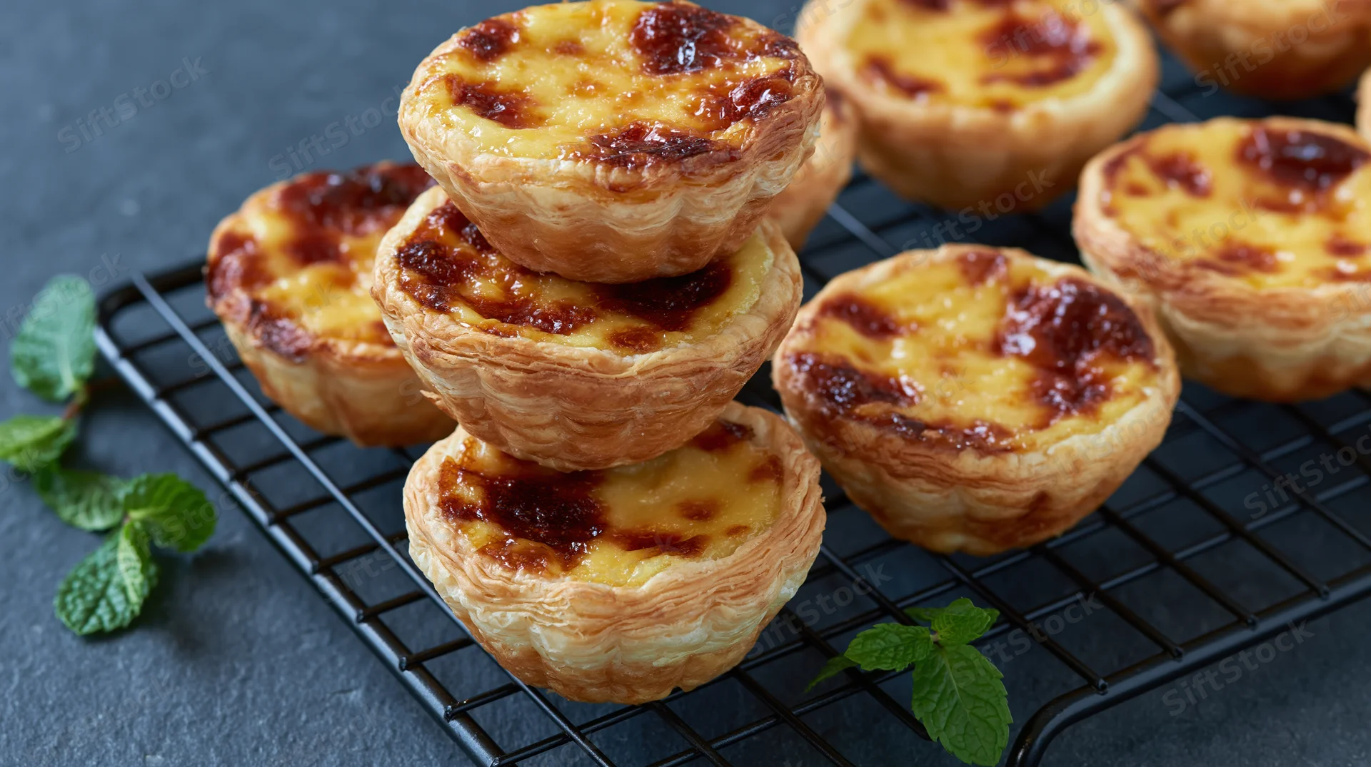 Portuguese egg tarts stacked on a cooling rack with mint garnish on dark slate surface