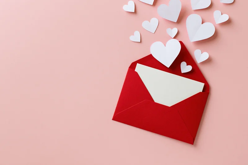 Red envelope open on pastel pink surface with white paper hearts emerging from it