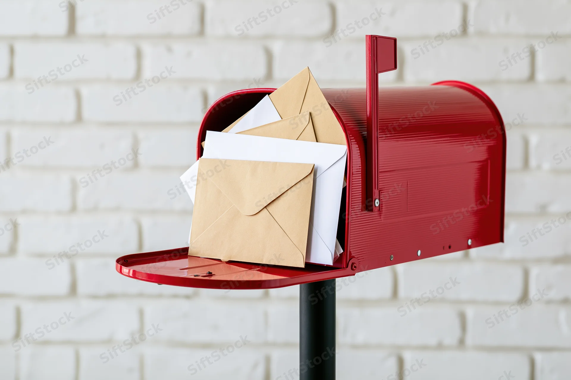 Red metal mailbox overflowing with letters and envelopes on black post against white brick wall