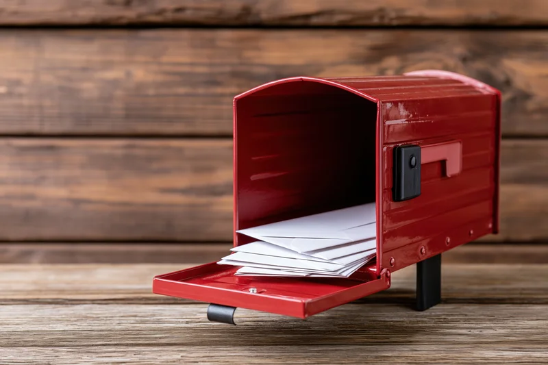 Red metal mailbox with open door revealing stack of white envelopes on wooden surface