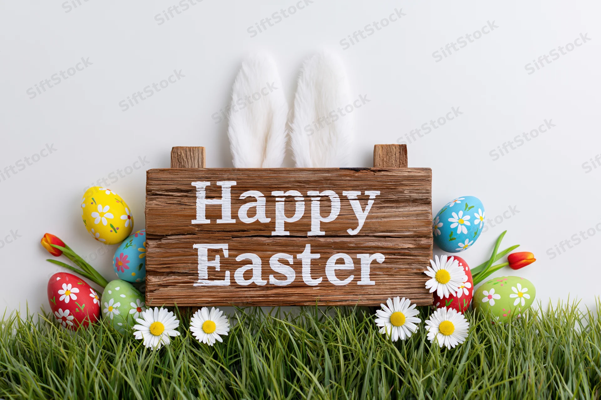 Rustic wooden sign reading happy easter propped in green grass with bunny ears and decorated eggs surrounded by daisies and tulips