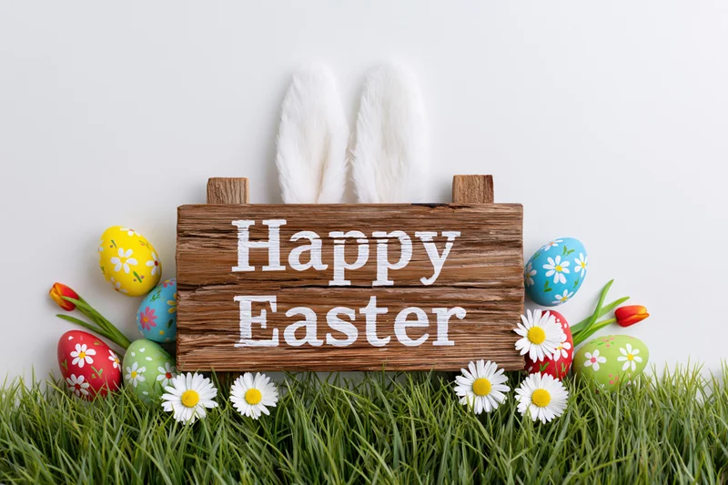 Rustic wooden sign reading happy easter propped in green grass with bunny ears and decorated eggs surrounded by daisies and tulips