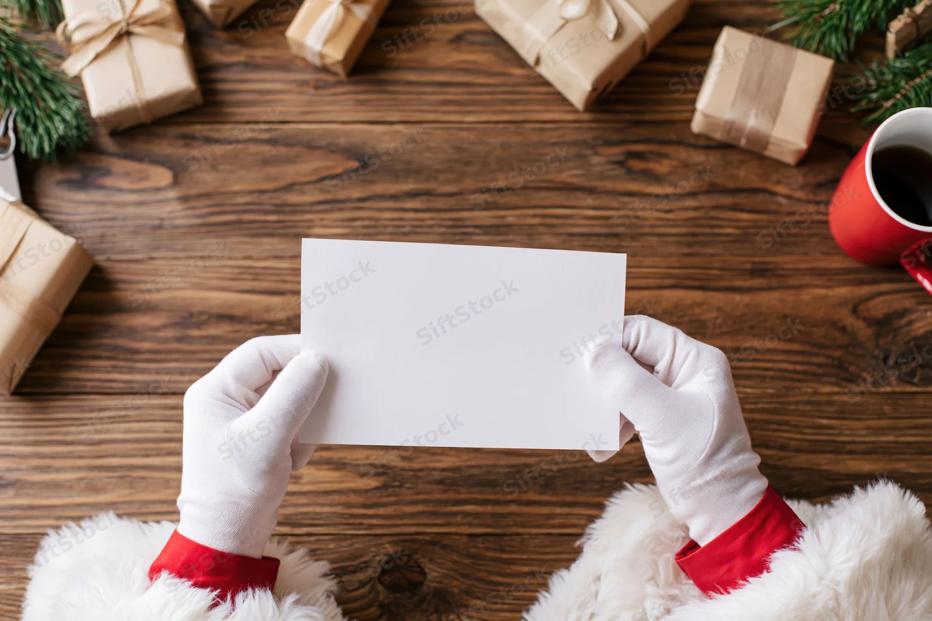 Santa's gloved hands holding a blank white card over a wooden table with wrapped gifts and pine branches