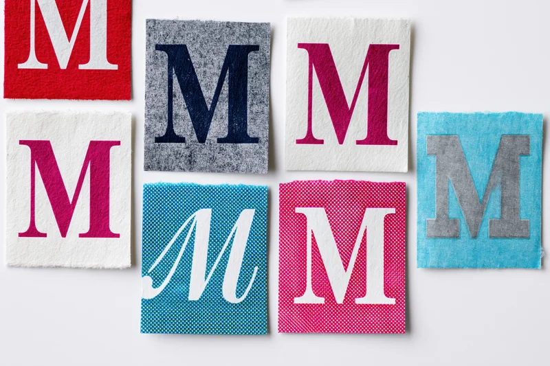Seven fabric squares each displaying the letter M in different colors and textures arranged on a white surface