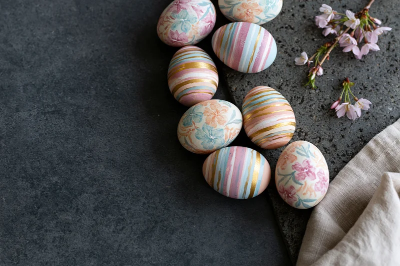 Decorated easter eggs with floral and striped patterns arranged on dark stone surface with cherry blossoms and linen cloth