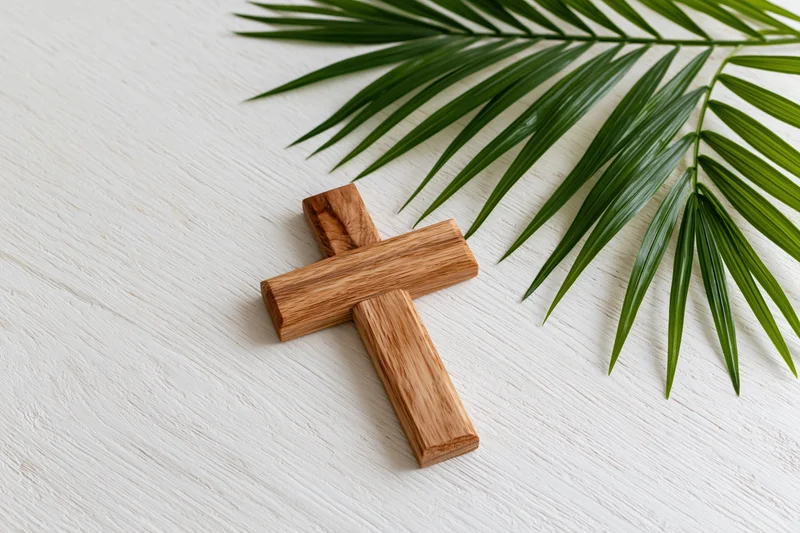 Wooden cross lying flat on white painted wood surface beside green palm leaf