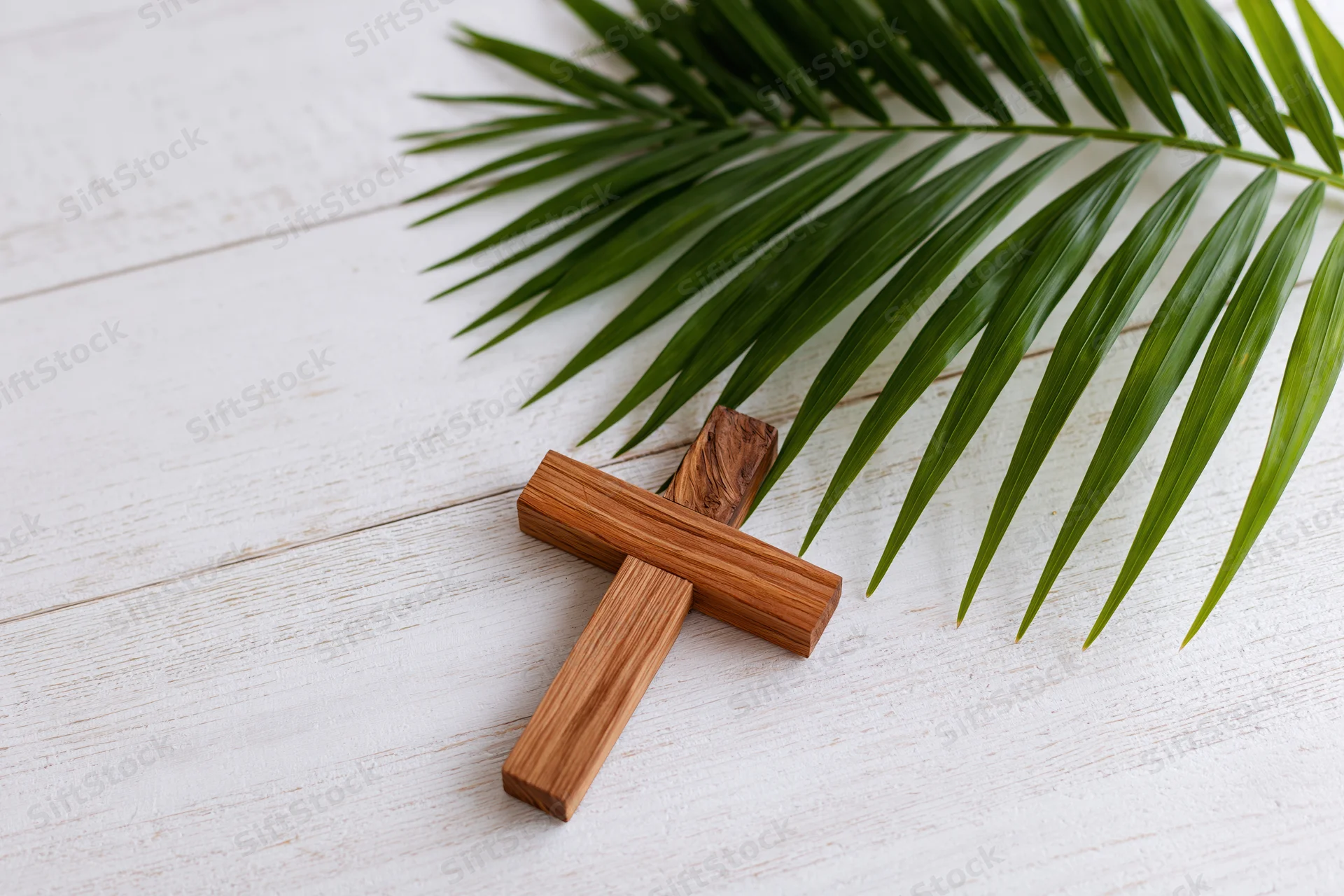 Wooden cross lying flat on white painted wood surface beside a green palm leaf