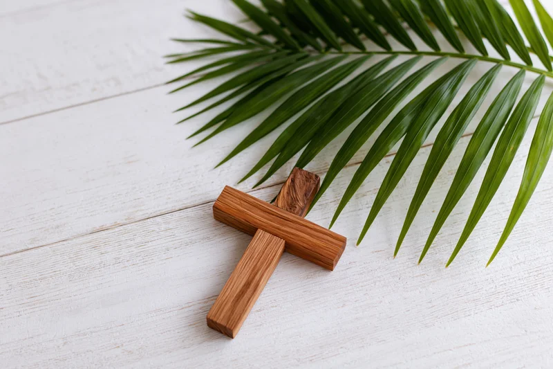 Wooden cross lying flat on white painted wood surface beside a green palm leaf