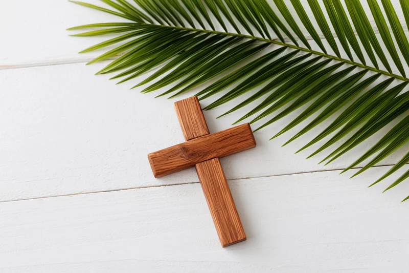 Wooden cross lying flat on white painted wood surface with palm leaf