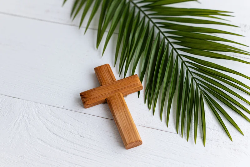 Wooden cross lying flat on white painted wood surface beside green palm leaf