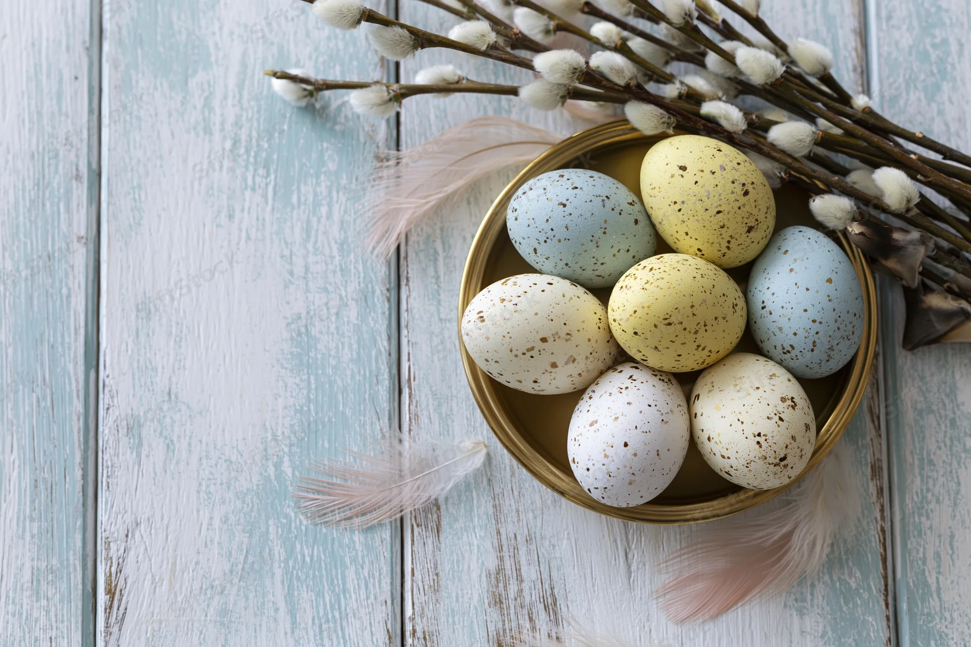 Speckled easter eggs in pastel colors arranged in a golden bowl with willow branches and feathers on a rustic wooden table