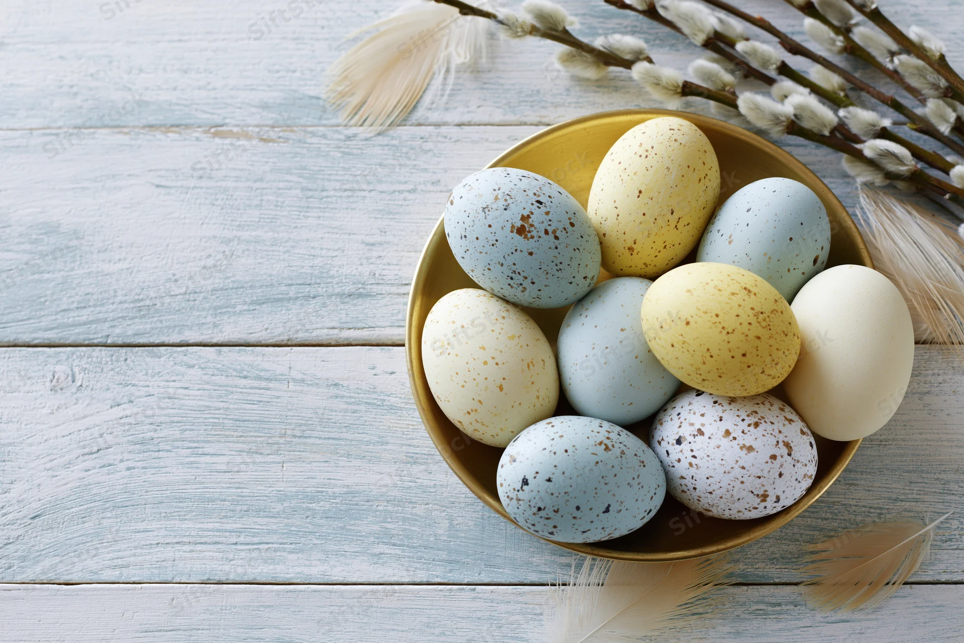 Speckled easter eggs in pastel colors arranged in a golden bowl on a wooden table with willow branches and feathers