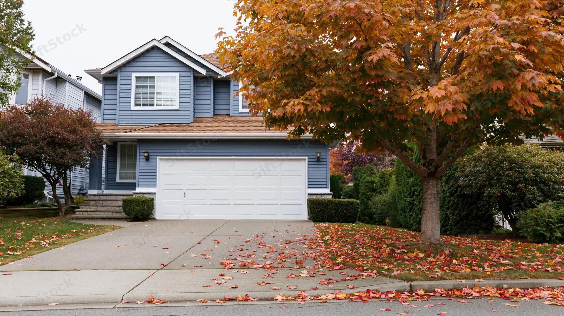 Suburban two-story house with blue-grey vinyl siding and white garage door, surrounded by autumn foliage and fallen leaves on the driveway and lawn