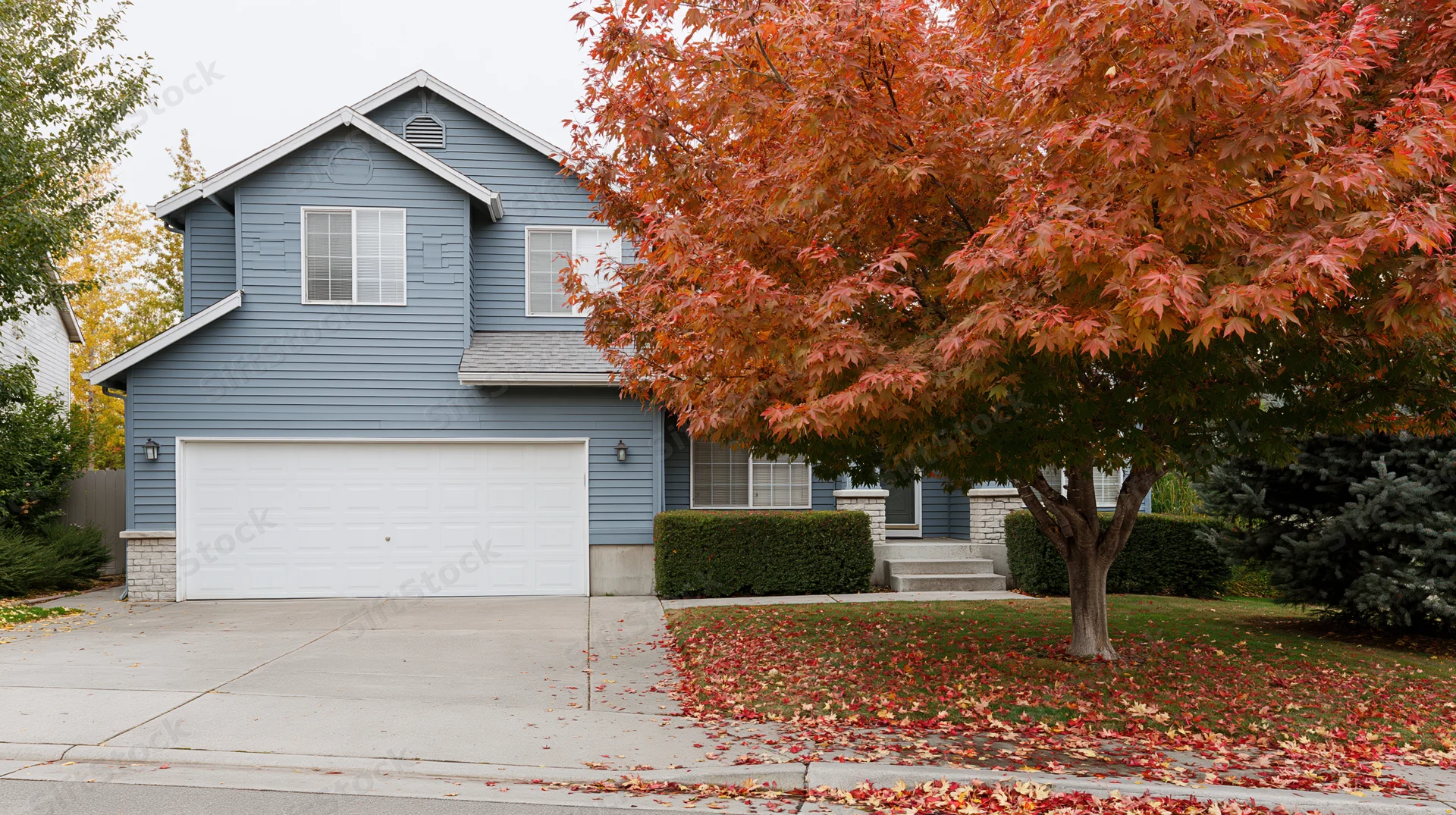 Suburban two-story house with blue-grey vinyl siding and white garage surrounded by autumn foliage and fallen leaves in front yard