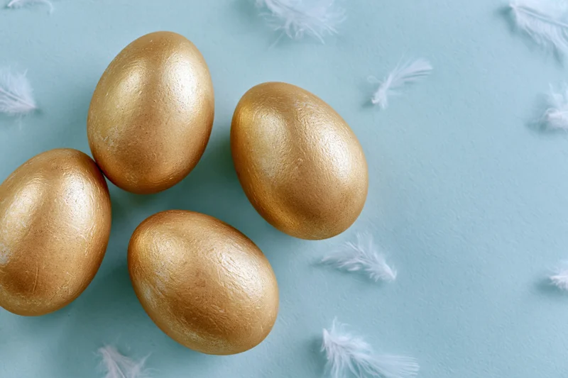 Four gold-painted easter eggs arranged on a blue surface with scattered white feathers