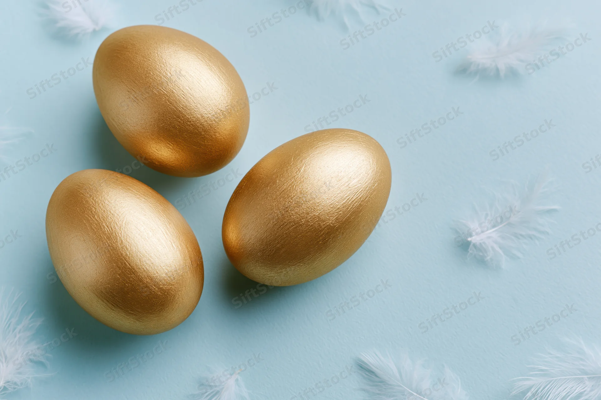 Three gold-painted easter eggs arranged on a light blue surface with scattered white feathers