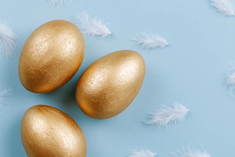 Three gold-painted easter eggs arranged loosely on a flat pastel blue surface with scattered white feathers