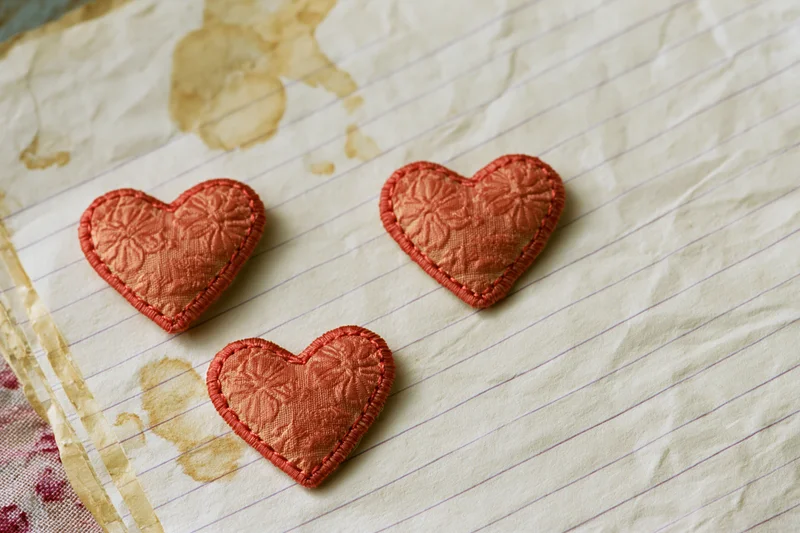 Three embroidered fabric hearts arranged on crumpled lined paper with coffee stains