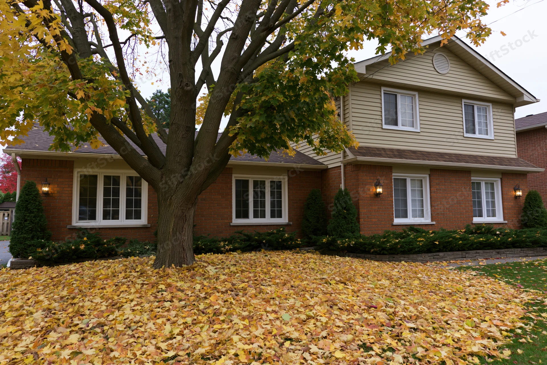 Two-story suburban house with brick and beige siding surrounded by fallen autumn leaves in front yard
