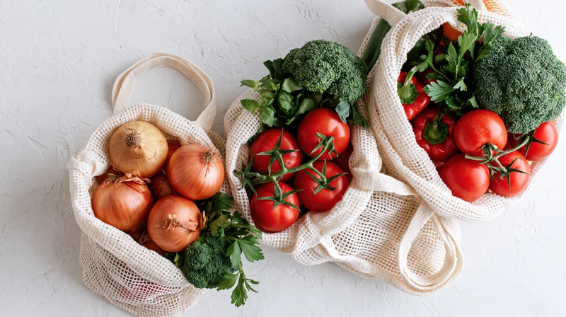 Three reusable mesh cotton tote bags filled with fresh vegetables arranged on a white surface
