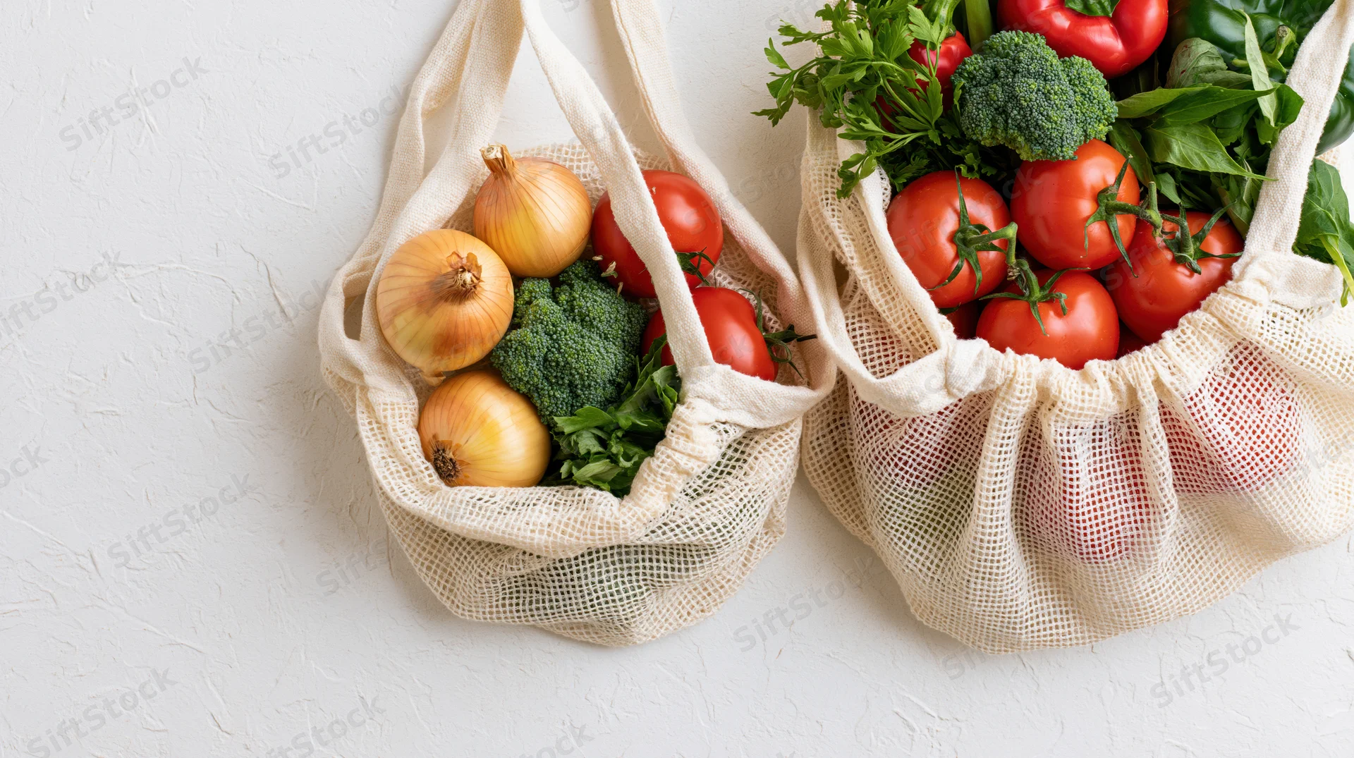Two reusable mesh cotton tote bags filled with fresh vegetables on a white surface