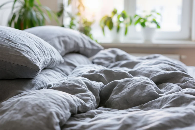 Unmade bed with rumpled grey linen duvet and stacked pillows in a bright bedroom with plants on the windowsill