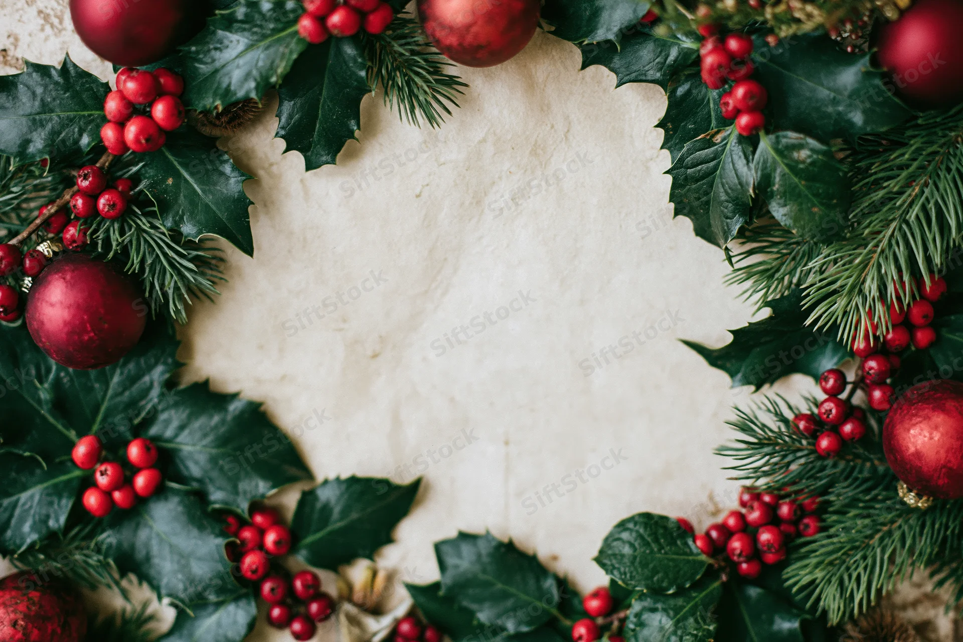 Circular christmas wreath made of holly leaves, red berries, pine needles and red ornaments framing a blank off-white center