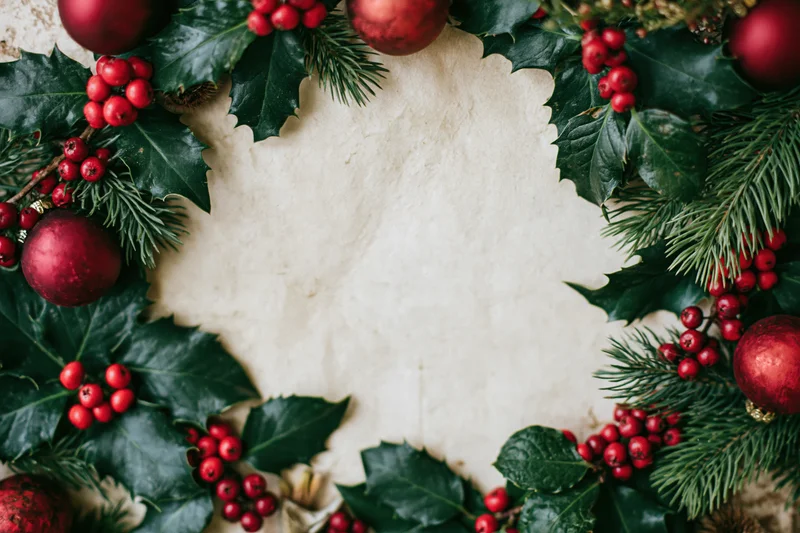 Circular christmas wreath made of holly leaves, red berries, pine needles and red ornaments framing a blank off-white center