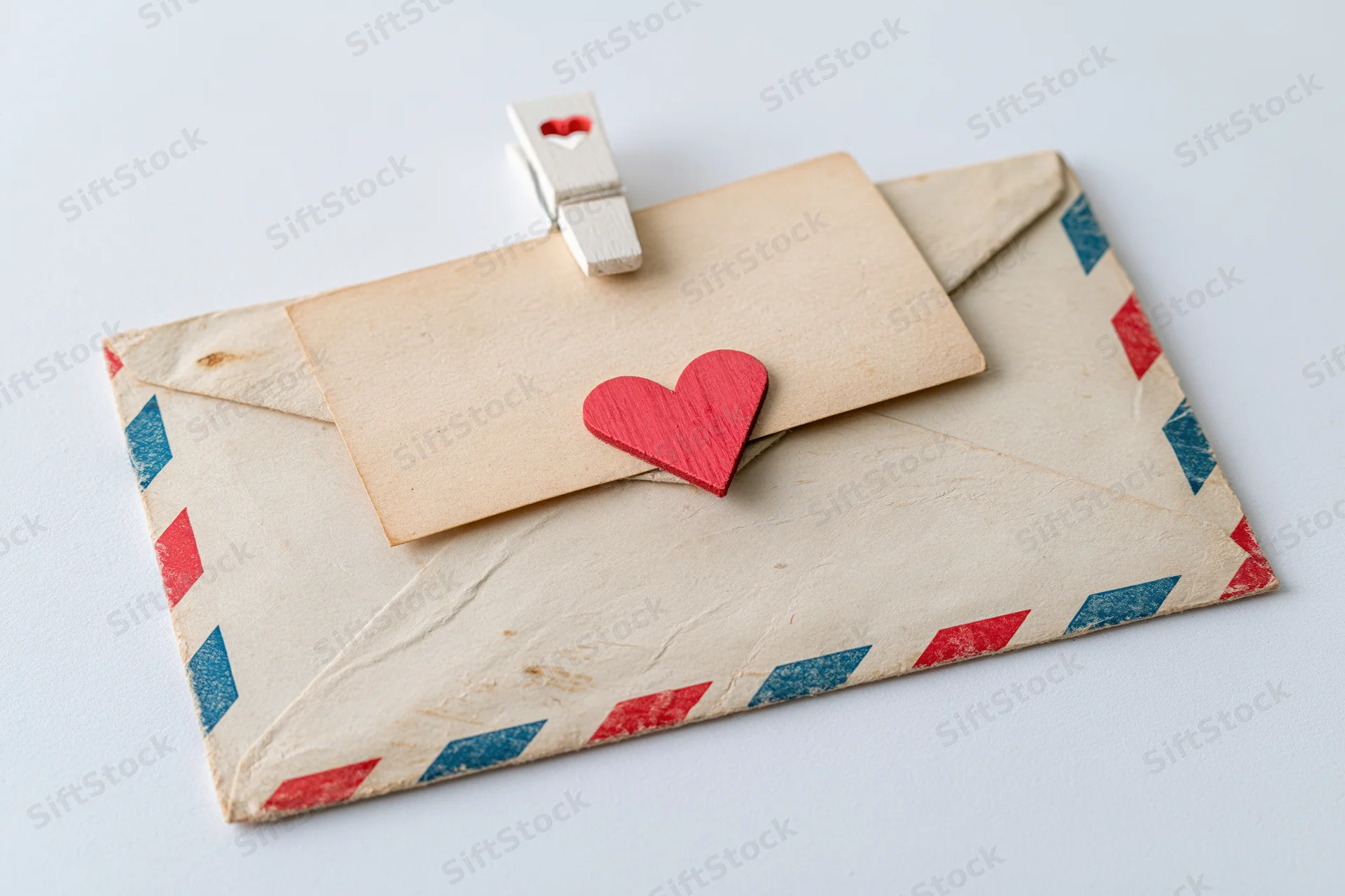 Vintage airmail envelope with red and blue stripes sealed with a wooden clothespin and a red heart decoration on white surface
