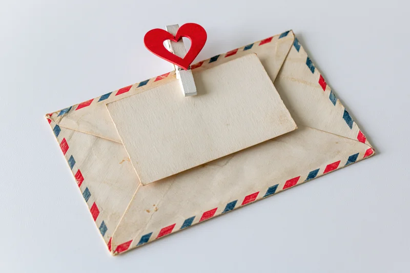 Vintage airmail envelope with red and blue stripes holding a blank card clipped by a red heart-shaped clothespin on white background