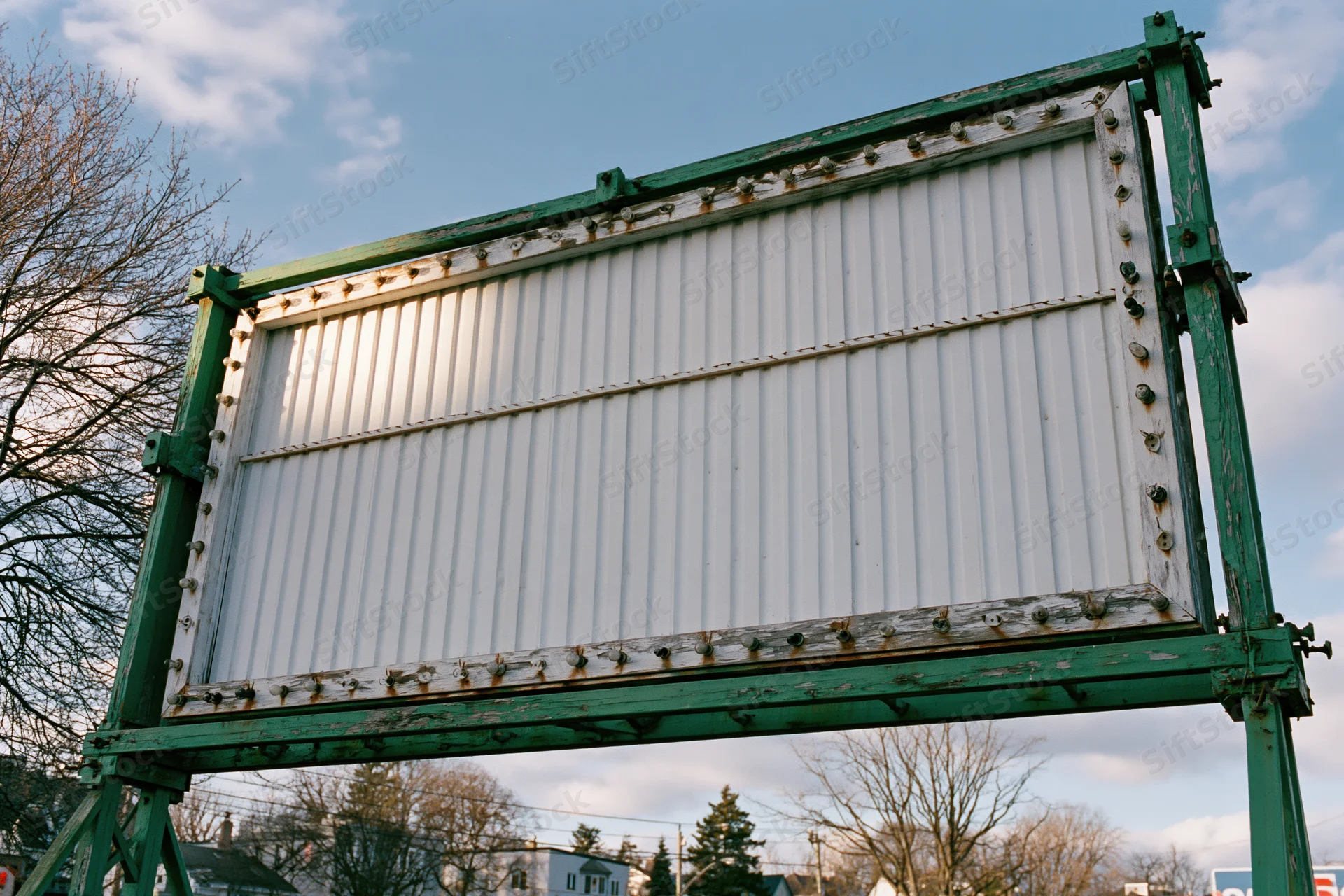 Weathered outdoor roadside marquee sign with empty white corrugated panels and green metal frame against a blue sky with bare trees in the background