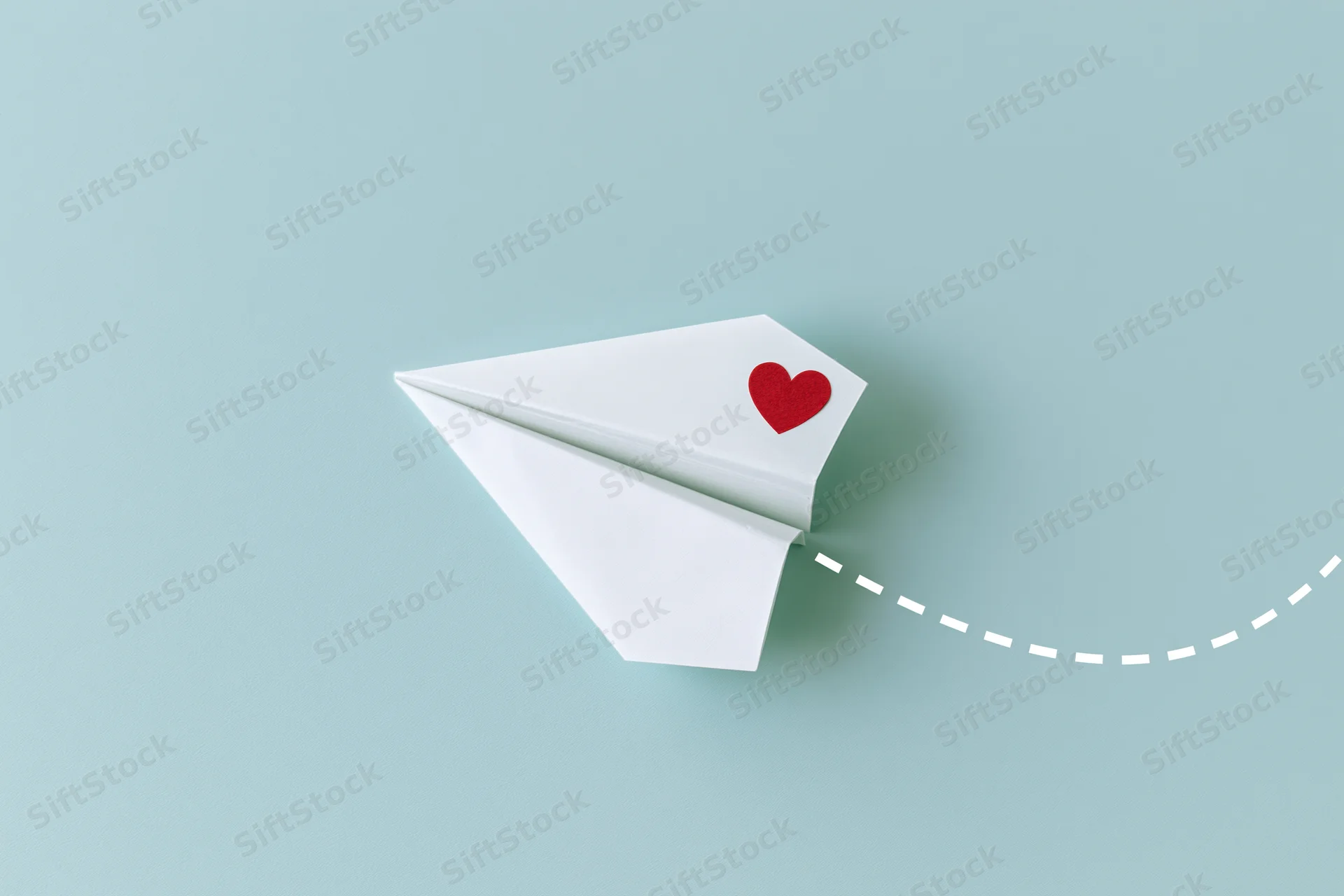 White origami paper airplane with red heart sticker flying on light blue background leaving dotted path
