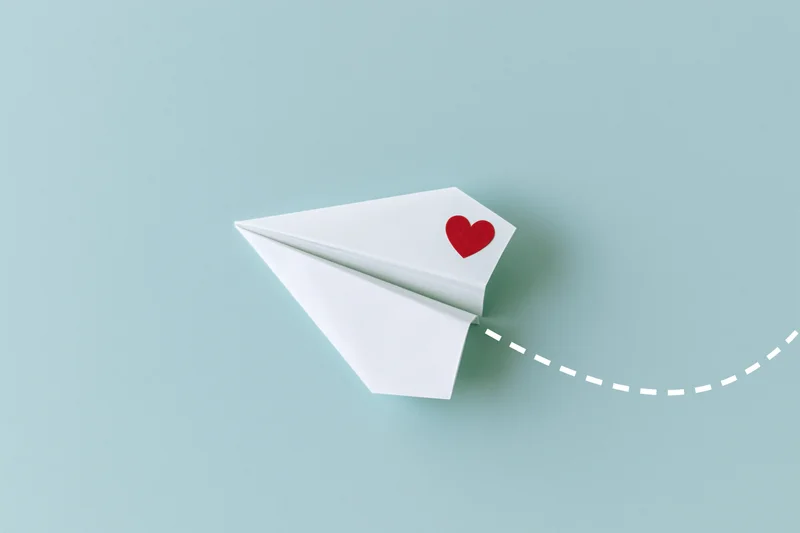 White origami paper airplane with red heart sticker flying on light blue background leaving dotted path