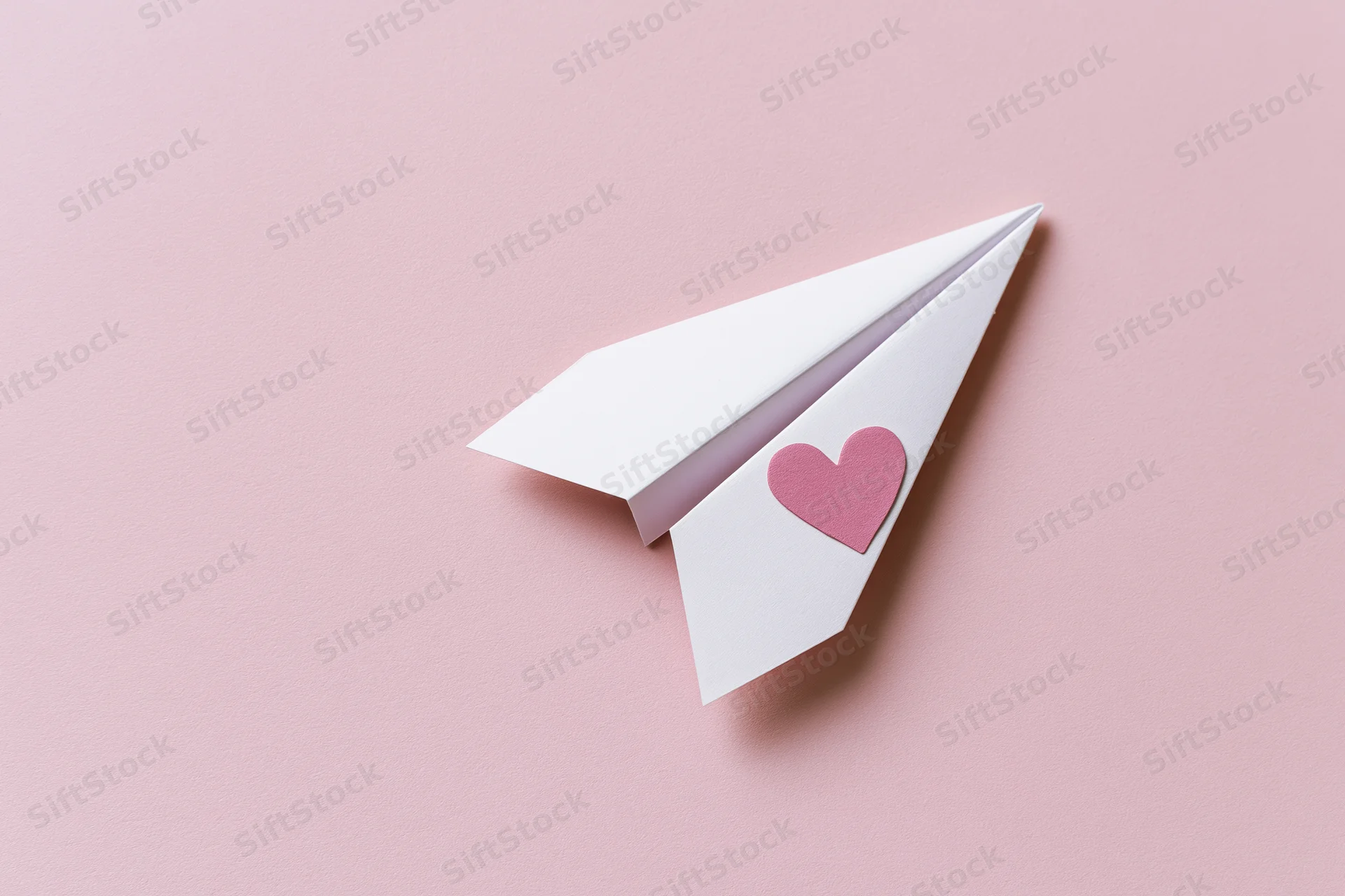 White paper airplane with pink heart sticker on pink surface