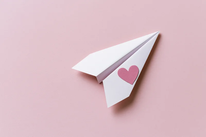White paper airplane with pink heart sticker on pink surface