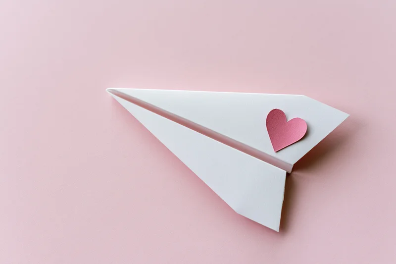 White paper airplane with pink heart sticker on pink surface