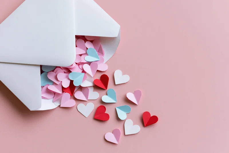 White envelope spilling colorful paper hearts onto pink surface